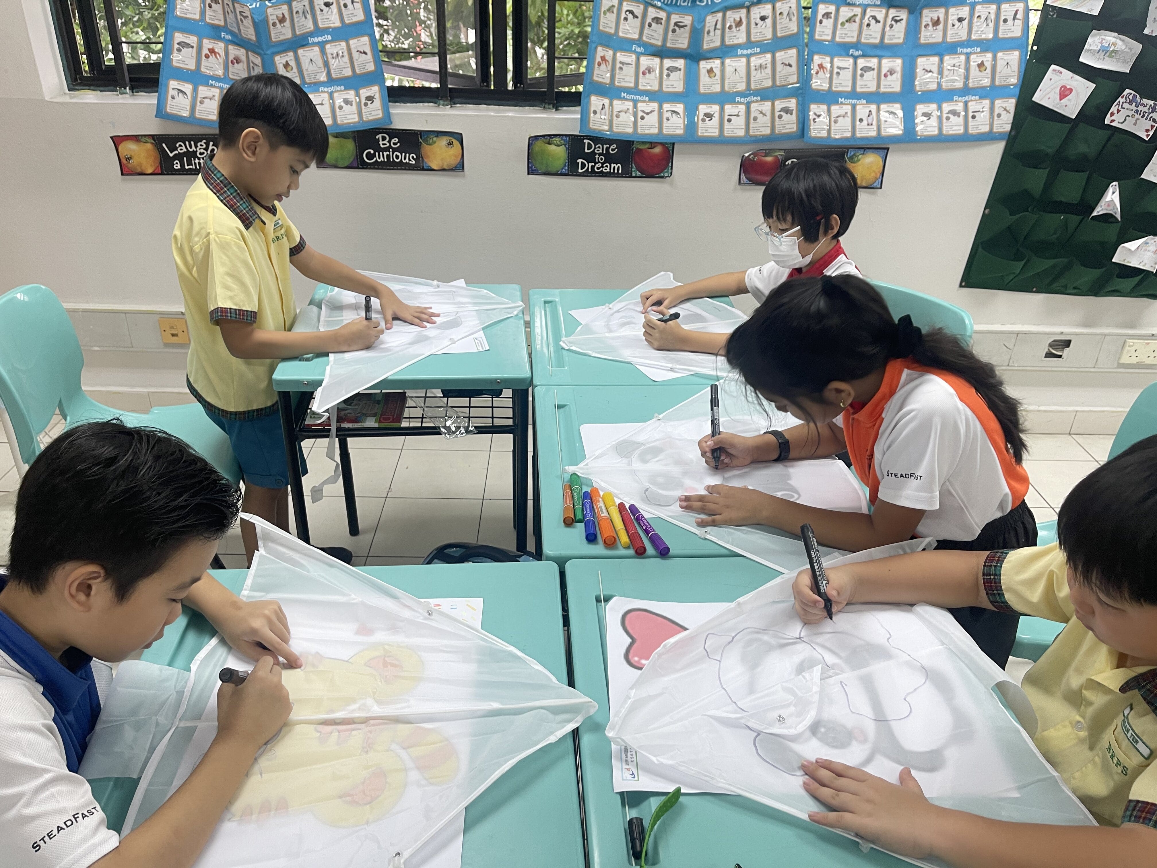 Four children in school uniforms draw with markers on white fabric kites at teal desks in a classroom.